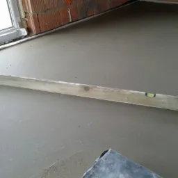 Świeżo wylana, szara posadzka wyr&oacute;wnywana poziomicą, widoczny fragment ściany z czerwonej cegły i fragment okna.