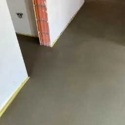 Świeżo wylana, gładka posadzka w surowym stanie, z widocznymi fragmentami ścian i żółtą taśmą dylatacyjną przy krawędziach, w trakcie prac wykończeniowych.