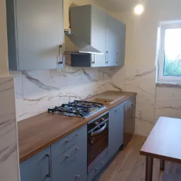 Jasnoszara kuchnia z drewnianym blatem, marmurowymi płytkami na ścianie i wiszącą żarówką bez klosza.