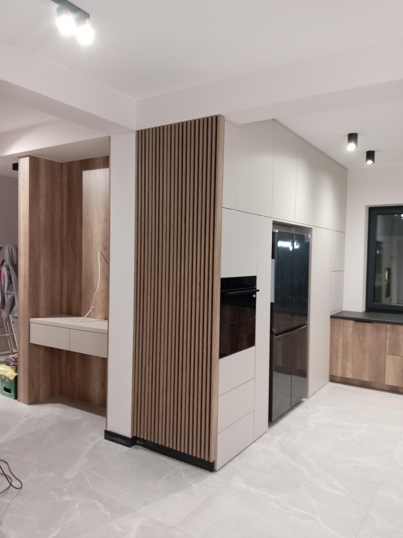 Nowoczesna kuchnia z białymi frontami, wbudowanym sprzętem AGD, drewnianymi akcentami w postaci pionowych lameli i blatu przy oknie, oraz marmurową podłogą.