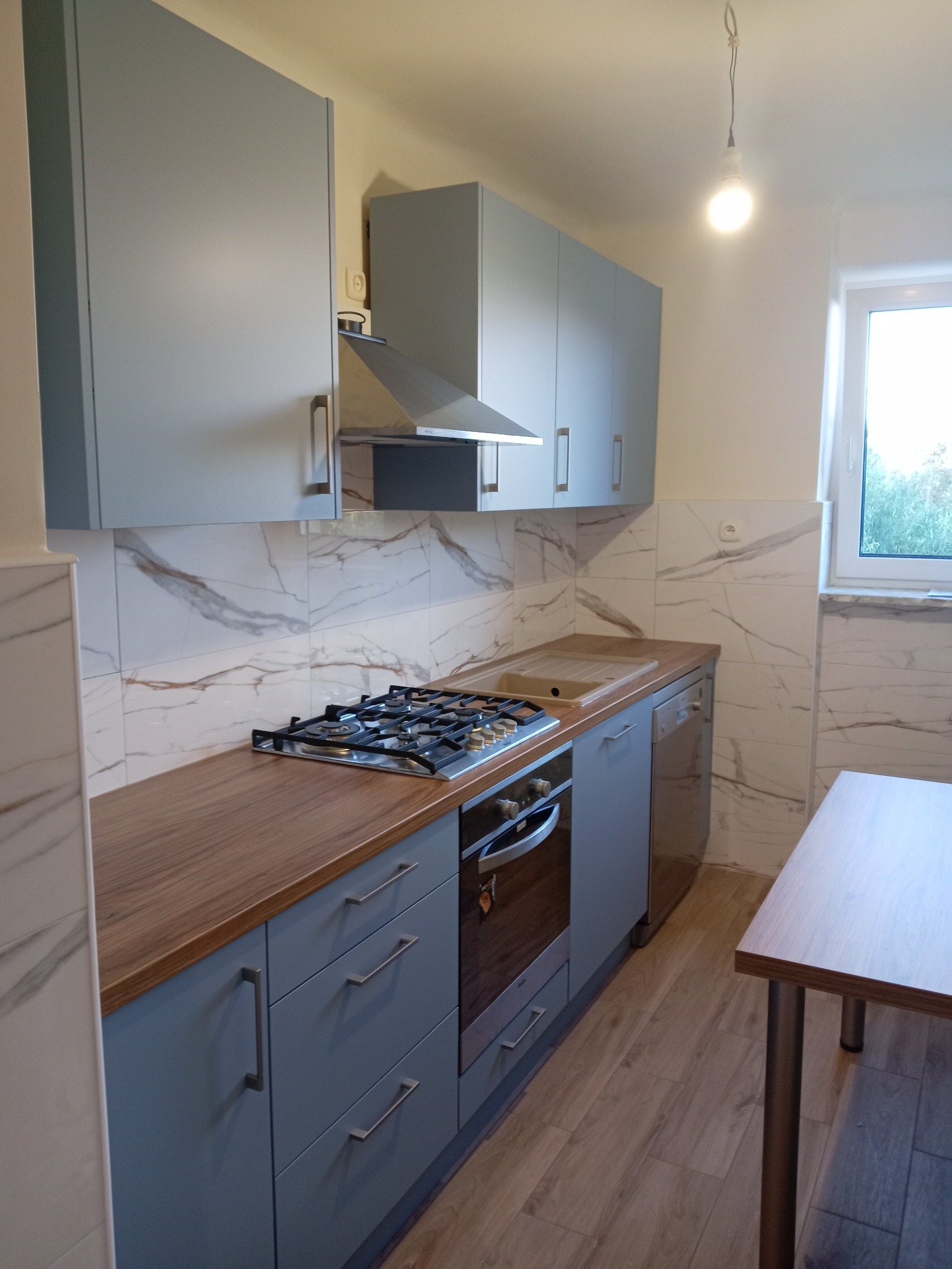 Jasnoszara kuchnia z drewnianym blatem, marmurowymi płytkami na ścianie i wiszącą żarówką bez klosza.