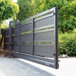 Nowoczesna, przesuwna brama wjazdowa w kolorze grafitowym, z poziomymi panelami, częściowo otwarta, na tle zieleni ogrodu i kostki brukowej.