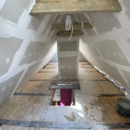 Poddasze w trakcie prac wykończeniowych: płyty gipsowo-kartonowe na ścianach ze szpachlowanymi łączeniami, surowa drewniana podłoga z odkrytym otworem, widoczne wiązki kabli i dwa wiadra z zaprawą.