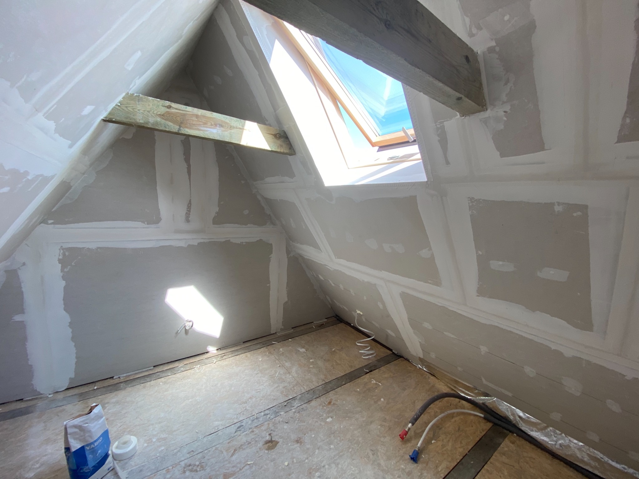 Poddasze w trakcie wykańczania płytami kartonowo-gipsowymi, widoczne szpachlowane łączenia, okno dachowe, drewniane belki konstrukcyjne, worek z gładzią i przewody na podłodze.