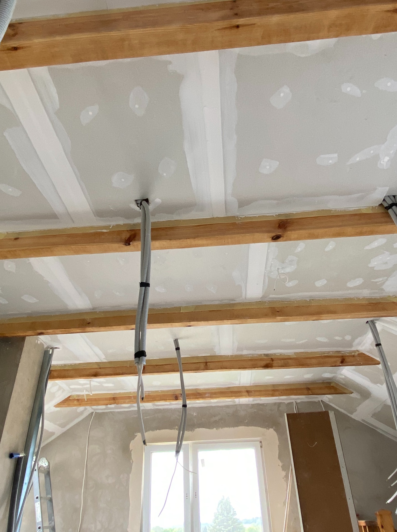 Poddasze w trakcie wykańczania: widoczne drewniane belki konstrukcyjne, płyty karton-gips z widocznymi łączeniami, wystające przewody elektryczne w peszlach, ściany w stanie surowym, drabina oparta...