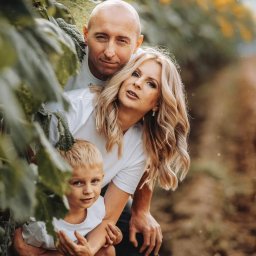 FotoArt Monika Górniak - Uśmiechnięta rodzina (ojciec, matka i syn) w białych koszulkach pozuje na tle pola słoneczników. Widoczny znak wodny fotografa w prawym górnym rogu.