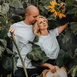FotoArt Monika Górniak - Para w polu słoneczników: blond kobieta trzyma kapelusz, mężczyzna obejmuje ją, oboje w białych koszulkach. Naturalne światło, zielone liście, ciepła atmosfera.