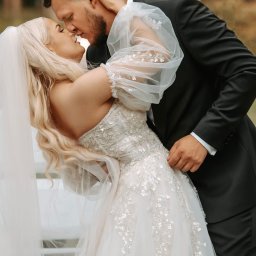 FotoArt Monika Górniak - Para młoda w objęciach podczas pocałunku. Panna młoda w sukni z delikatnymi zdobieniami, pan młody w czarnym garniturze. Romantyczna atmosfera ślubna.