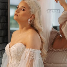 FotoArt Monika Górniak - Panna młoda w sukni ślubnej, z blond włosami i błyszczącymi kolczykami, podczas zakładania welonu przed lustrem. Delikatny makijaż i zdobiona suknia.