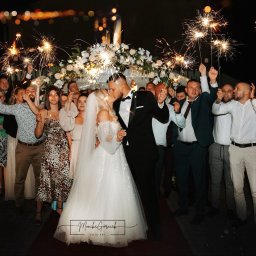 FotoArt Monika Górniak - Pocałunek Młodej Pary otoczonej gośćmi z zimnymi ogniami na tle kwiecistej dekoracji ślubnej, nocna sceneria z rozmytymi światłami w tle.