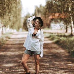 FotoArt Monika Górniak - Portret kobiety w kapeluszu i rozpiętej koszuli, na leśnej drodze. Uśmiechnięta modelka pozuje w swobodnej stylizacji, z logo fotografa w tle. Naturalne światło, plener.