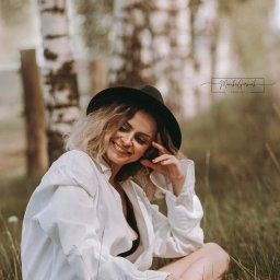 FotoArt Monika Górniak - Portret uśmiechniętej kobiety w kapeluszu, siedzącej na trawie w lesie brzozowym, w luźnej białej koszuli, z delikatnym makijażem i pomalowanymi paznokciami.