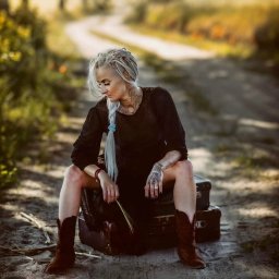 FotoArt Monika Górniak - Kobieta z blond warkoczami i tatuażami siedzi na walizkach w brązowych kowbojkach na polnej drodze. W ręku trzyma źdźbło trawy. Ujęcie w plenerze, w tle rozmyta droga.