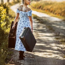 FotoArt Monika Górniak - Kobieta z tatuażem na plecach, w kwiecistej sukience, idzie polną drogą, niosąc dwie walizki w stylu vintage. Ujęcie z tyłu, w ciepłym świetle.