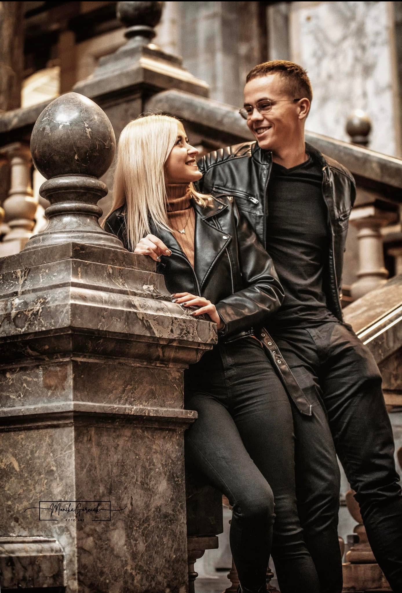 Uśmiechnięta para w skórzanych kurtkach pozuje na marmurowych schodach. Kobieta opiera się o balustradę, mężczyzna siedzi obok. Stonowana kolorystyka, portret.