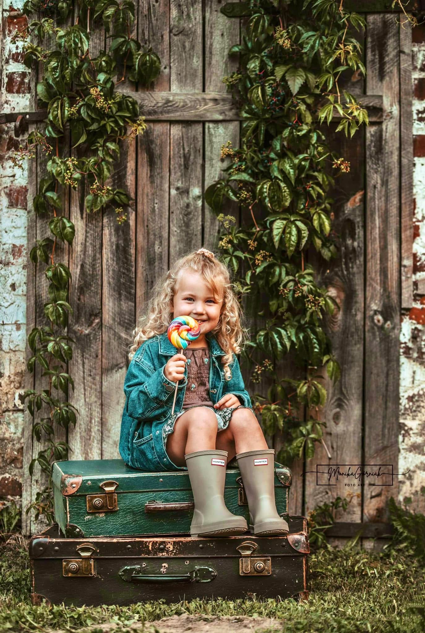 Uśmiechnięta dziewczynka z lizakiem siedzi na walizkach w gumowcach Hunter, na tle drewnianych drzwi oplecionych bluszczem. Sesja w plenerze w rustykalnym stylu.