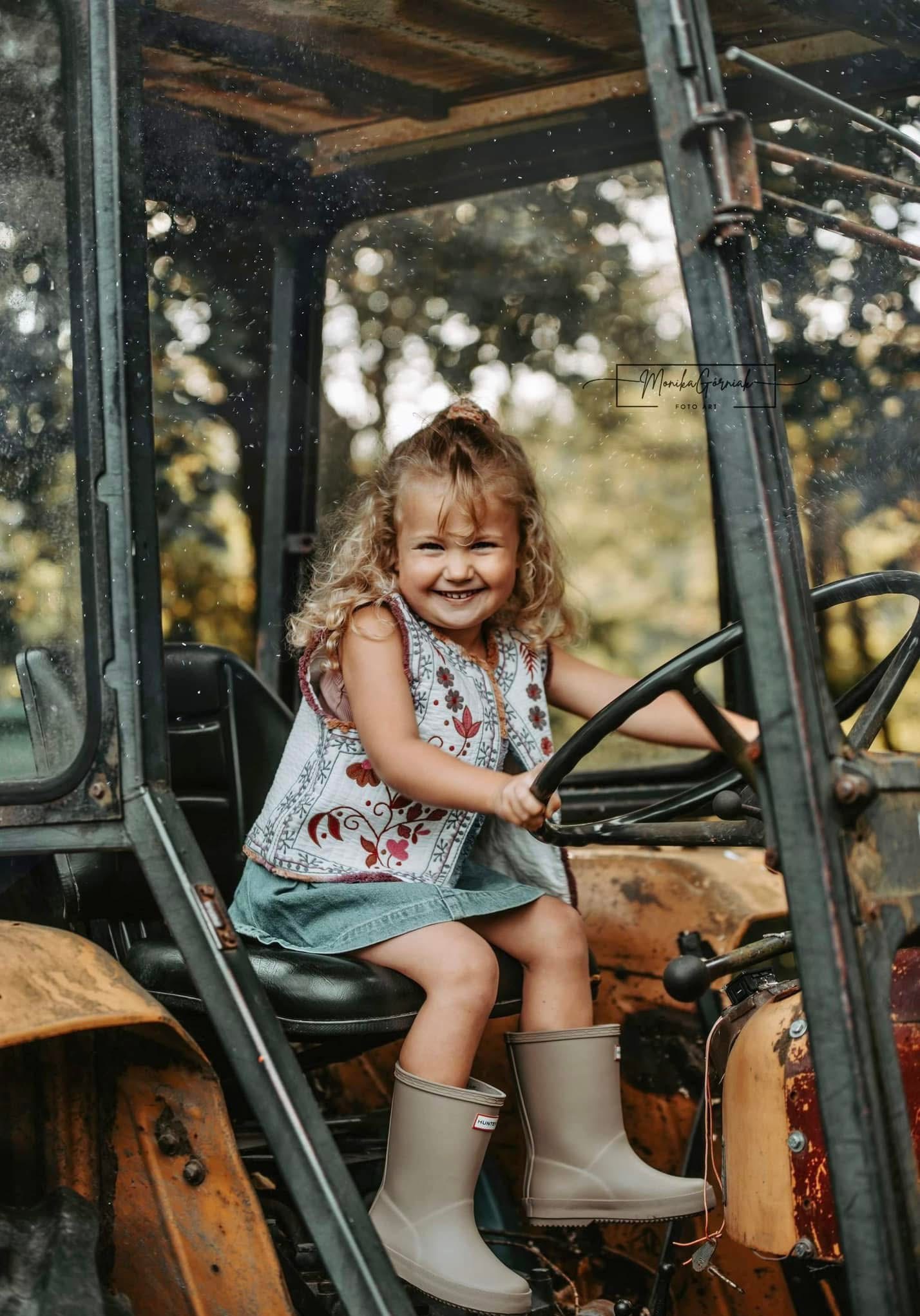 Uśmiechnięta dziewczynka w kwiecistej kamizelce siedzi za kierownicą starego traktora, w gumowych kaloszach. Rozmyte tło drzew. Ciepłe, naturalne światło.