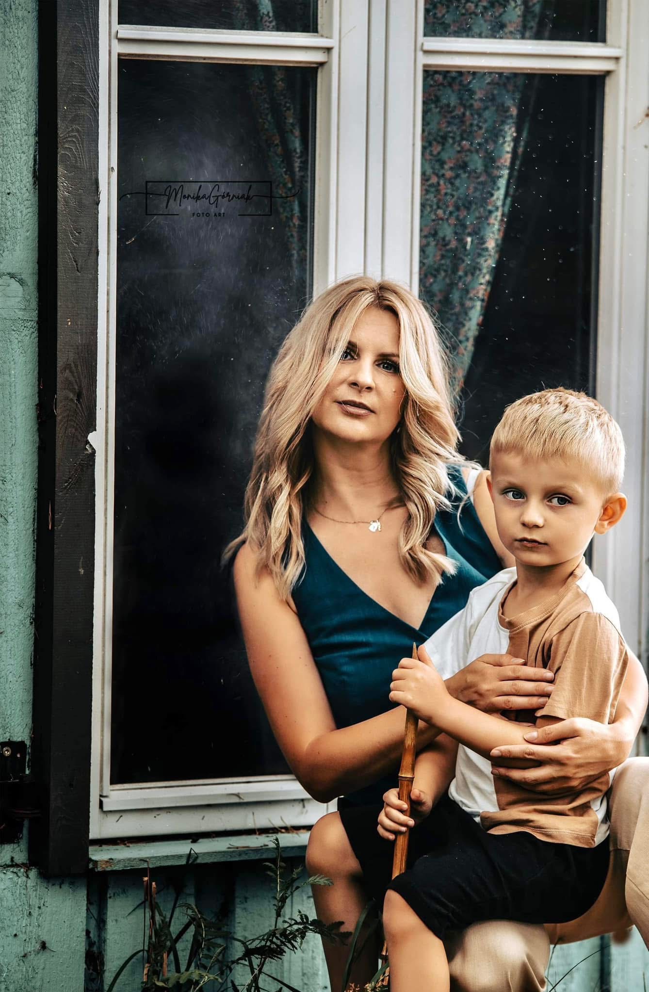 Portret mamy z synkiem w Kołobrzegu na tle okna. Kobieta w zielonej sukience, chłopiec trzyma bambusowy kijek. Naturalne światło, spokojna atmosfera.