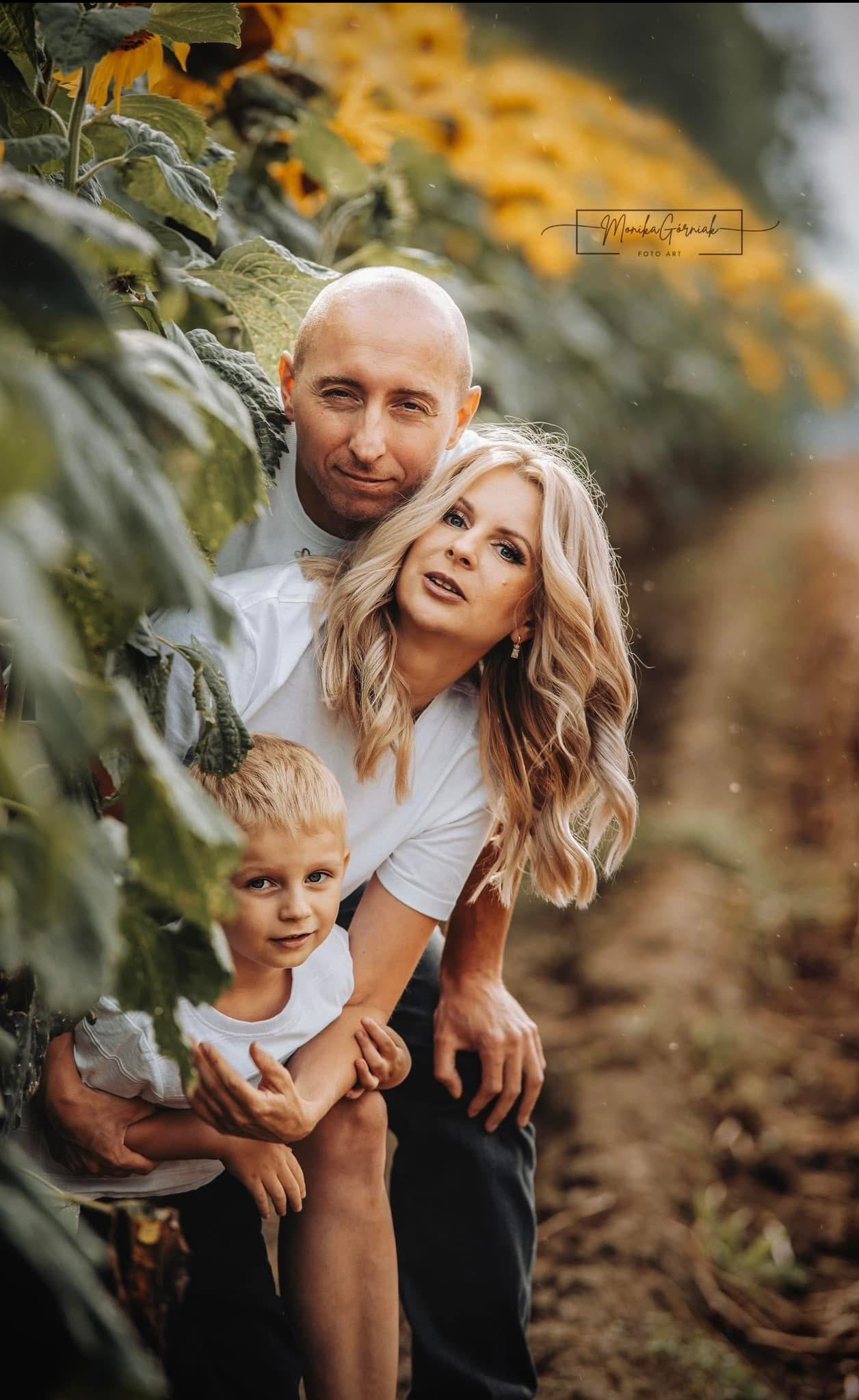 Uśmiechnięta rodzina (ojciec, matka i syn) w białych koszulkach pozuje na tle pola słoneczników. Widoczny znak wodny fotografa w prawym górnym rogu.