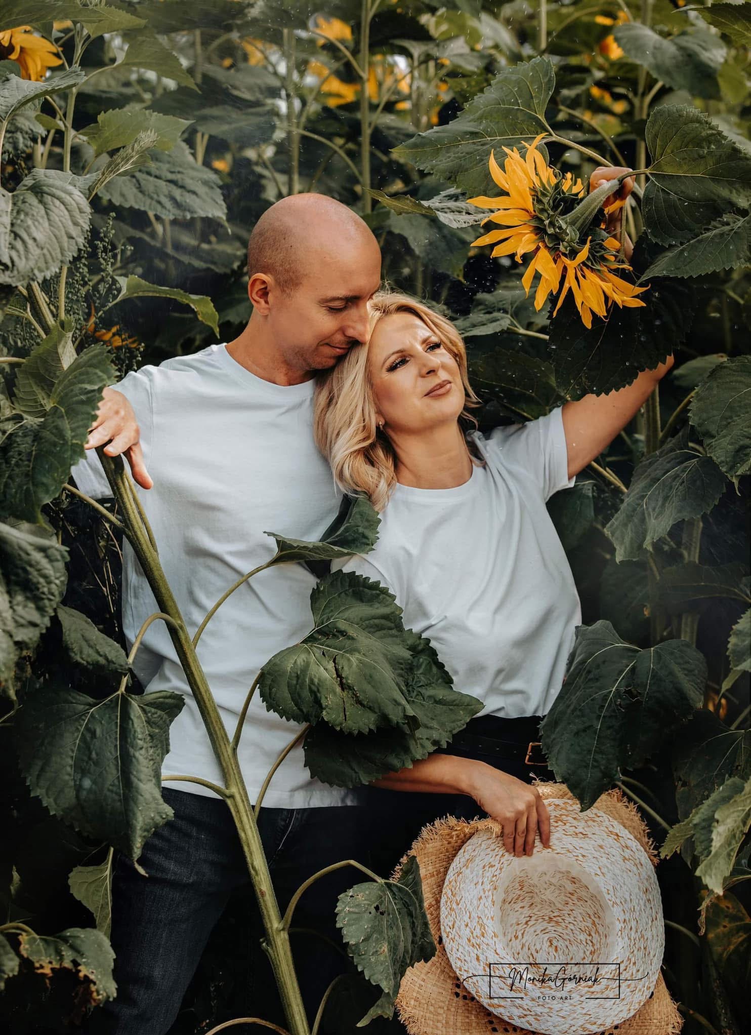 Para w polu słoneczników: blond kobieta trzyma kapelusz, mężczyzna obejmuje ją, oboje w białych koszulkach. Naturalne światło, zielone liście, ciepła atmosfera.