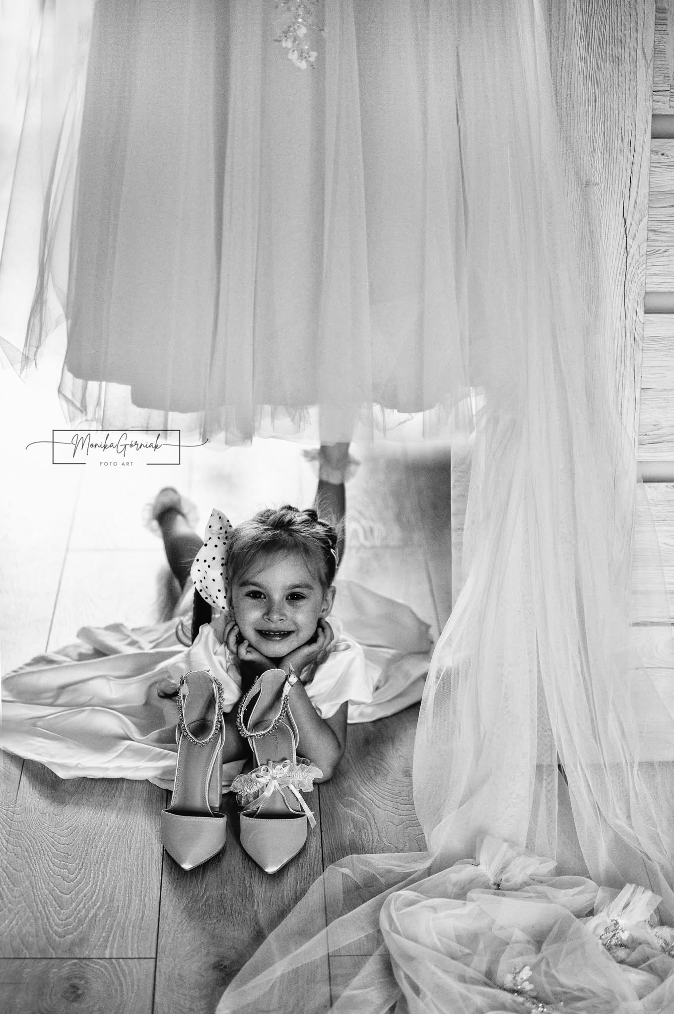 Uśmiechnięta dziewczynka z kokardą leży na podłodze obok butów ślubnych, pod suknią ślubną. Czarno-biała fotografia z subtelnym oświetleniem.