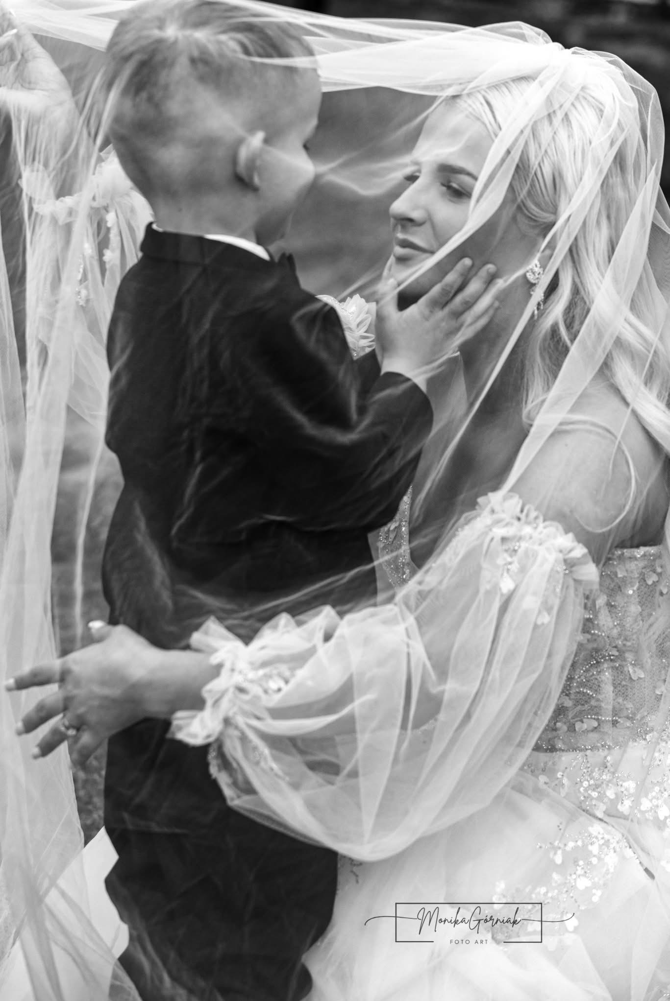 Czarno-białe ujęcie panny młodej w welonie, delikatnie dotykanej przez małego chłopca w eleganckim ubraniu. Romantyczna atmosfera, subtelne światło.