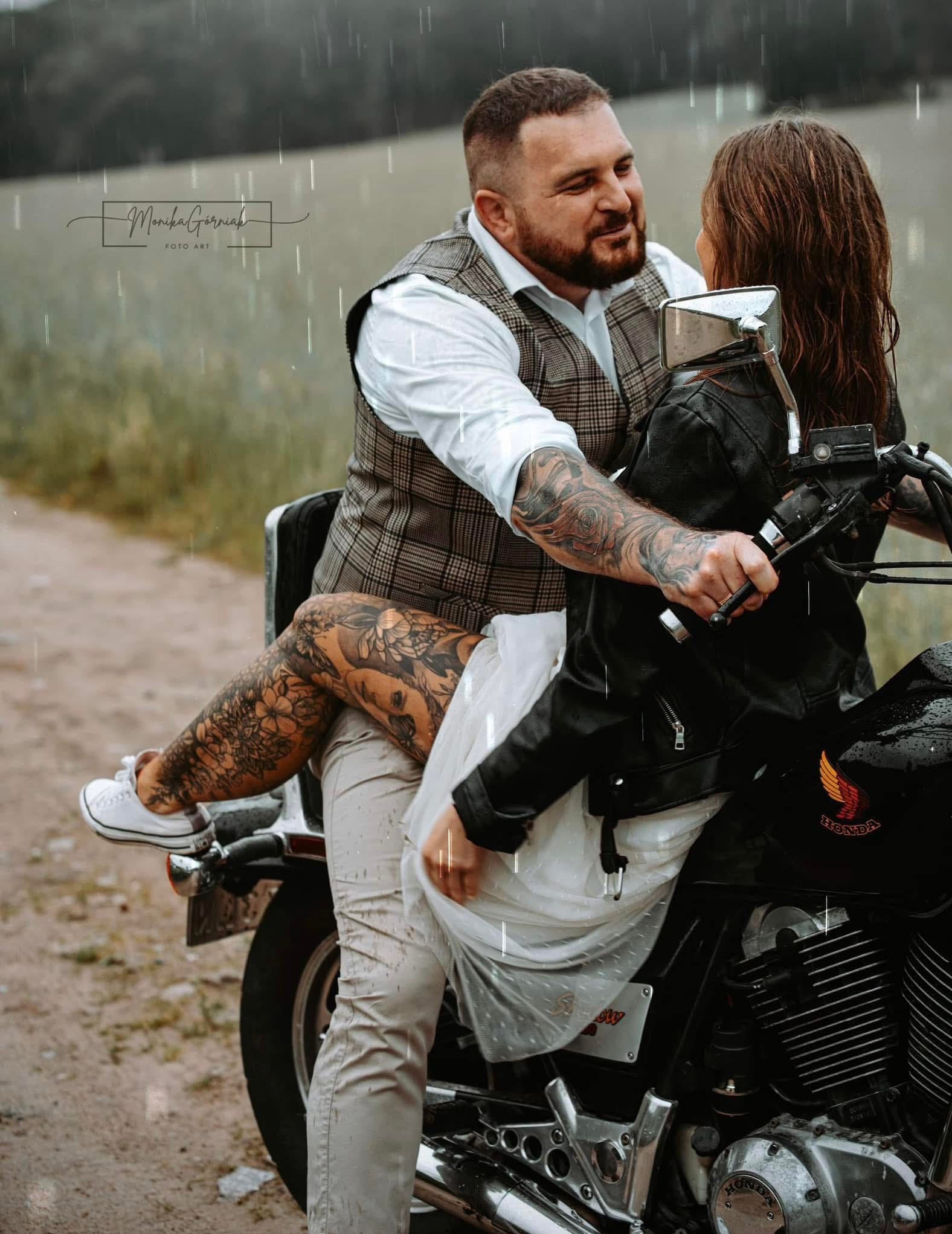 Zakochana para na motocyklu Honda Shadow w deszczu. Mężczyzna w kraciastej kamizelce obejmuje kobietę w białej sukience i czarnej kurtce. Widoczne tatuaże na ciele.