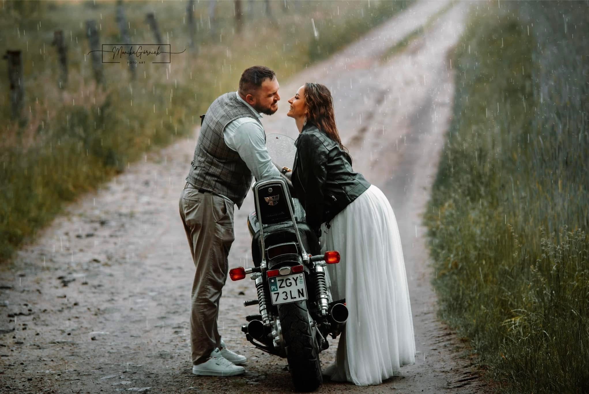 Zakochana para w deszczu, oparta o motocykl. Ona w białej sukni i czarnej kurtce, on w kamizelce i spodniach. Romantyczna sesja na polnej drodze.