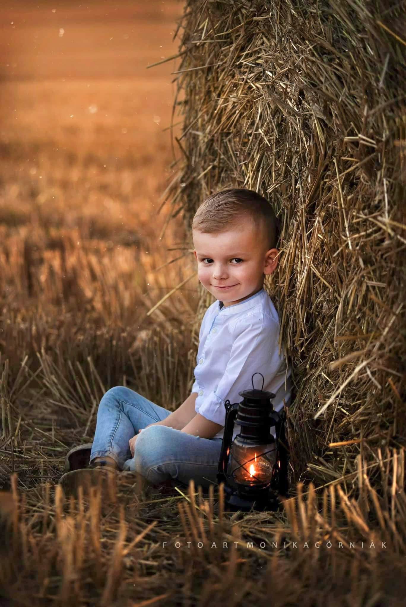 Uśmiechnięty chłopiec w jeansach siedzi obok stogu siana, trzymając zapaloną, czarną latarnię. Ciepłe, naturalne światło. Na dole widoczny podpis fotografa.