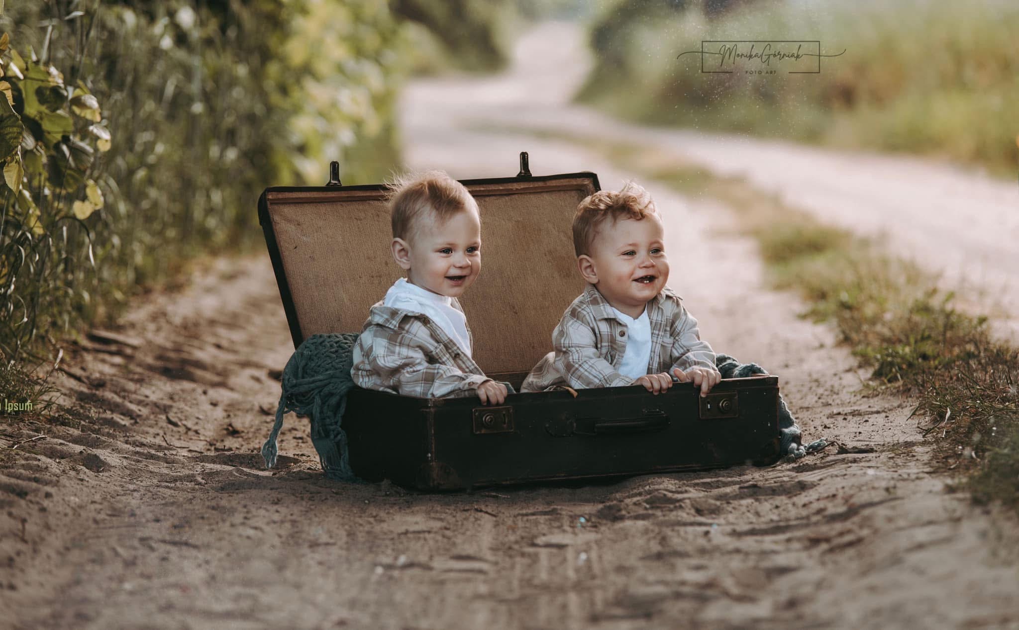 Uśmiechnięte bliźniaki w brązowej walizce na polnej drodze, w tle zieleń. Sesja w plenerze z uroczymi chłopcami w koszulach w kratę.