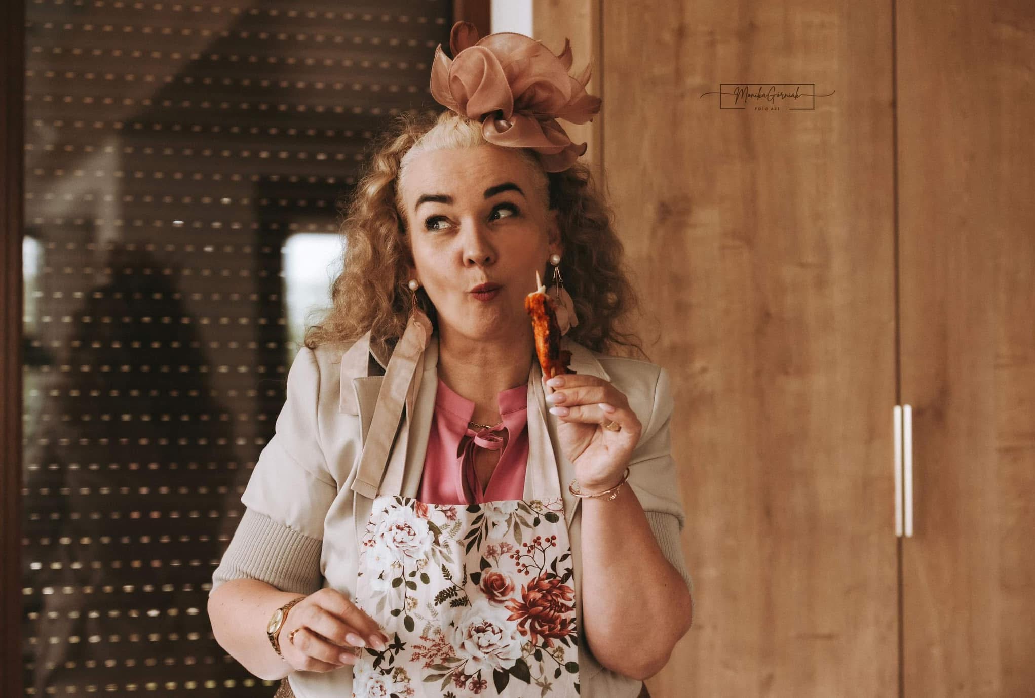 Kobieta z kapeluszem i w kwiecistym fartuchu, trzymająca przekąskę na patyku, patrzy w lewo na tle drewnianej ściany i okna z roletami.