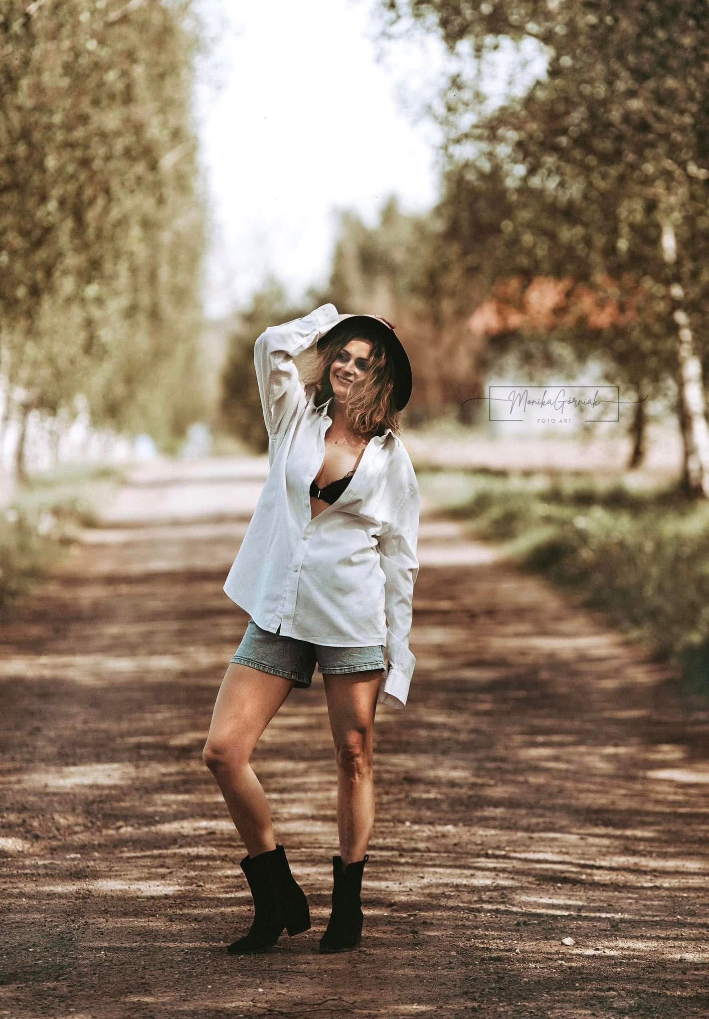 Portret kobiety w kapeluszu i rozpiętej koszuli, na leśnej drodze. Uśmiechnięta modelka pozuje w swobodnej stylizacji, z logo fotografa w tle. Naturalne światło, plener.