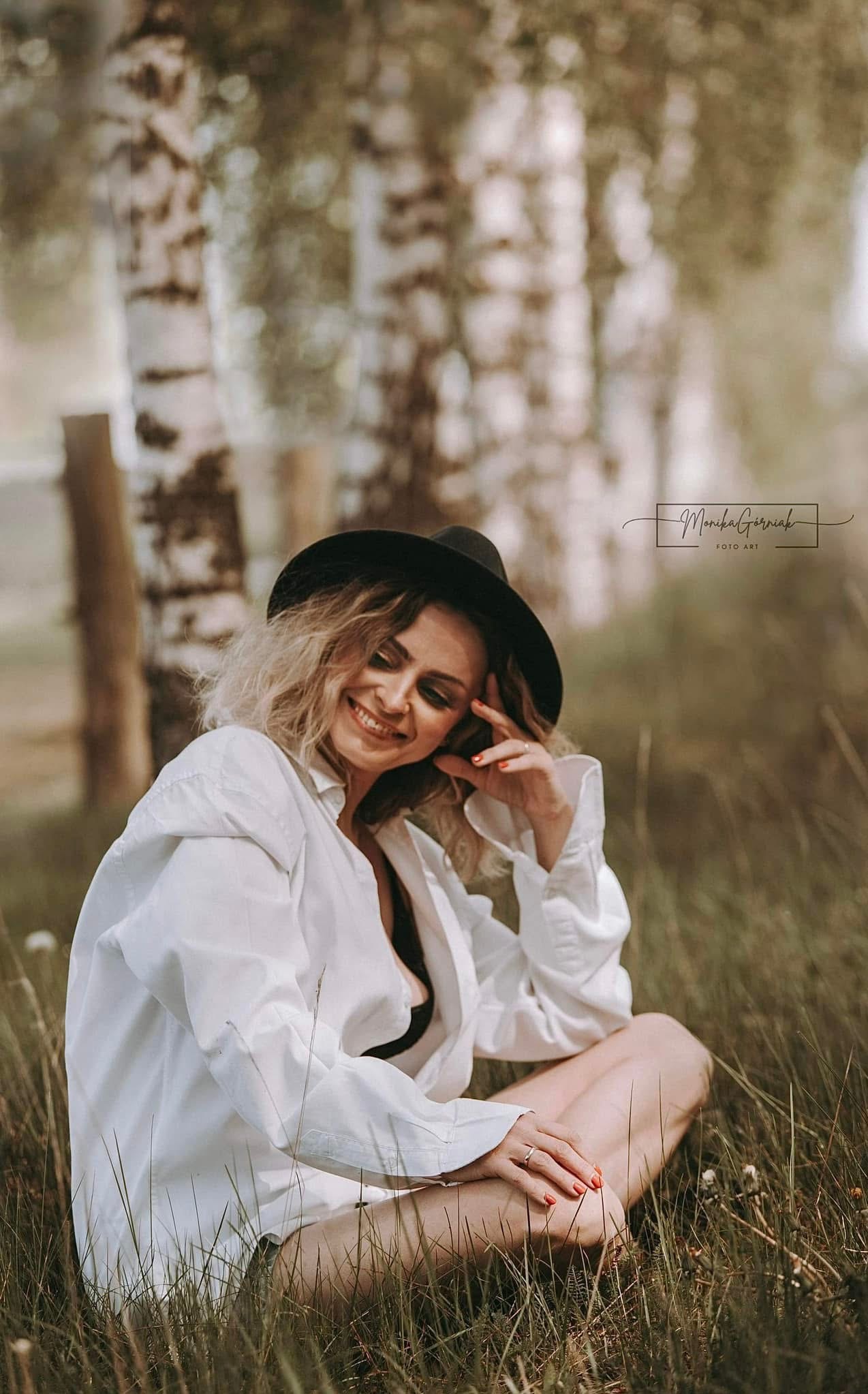 Portret uśmiechniętej kobiety w kapeluszu, siedzącej na trawie w lesie brzozowym, w luźnej białej koszuli, z delikatnym makijażem i pomalowanymi paznokciami.