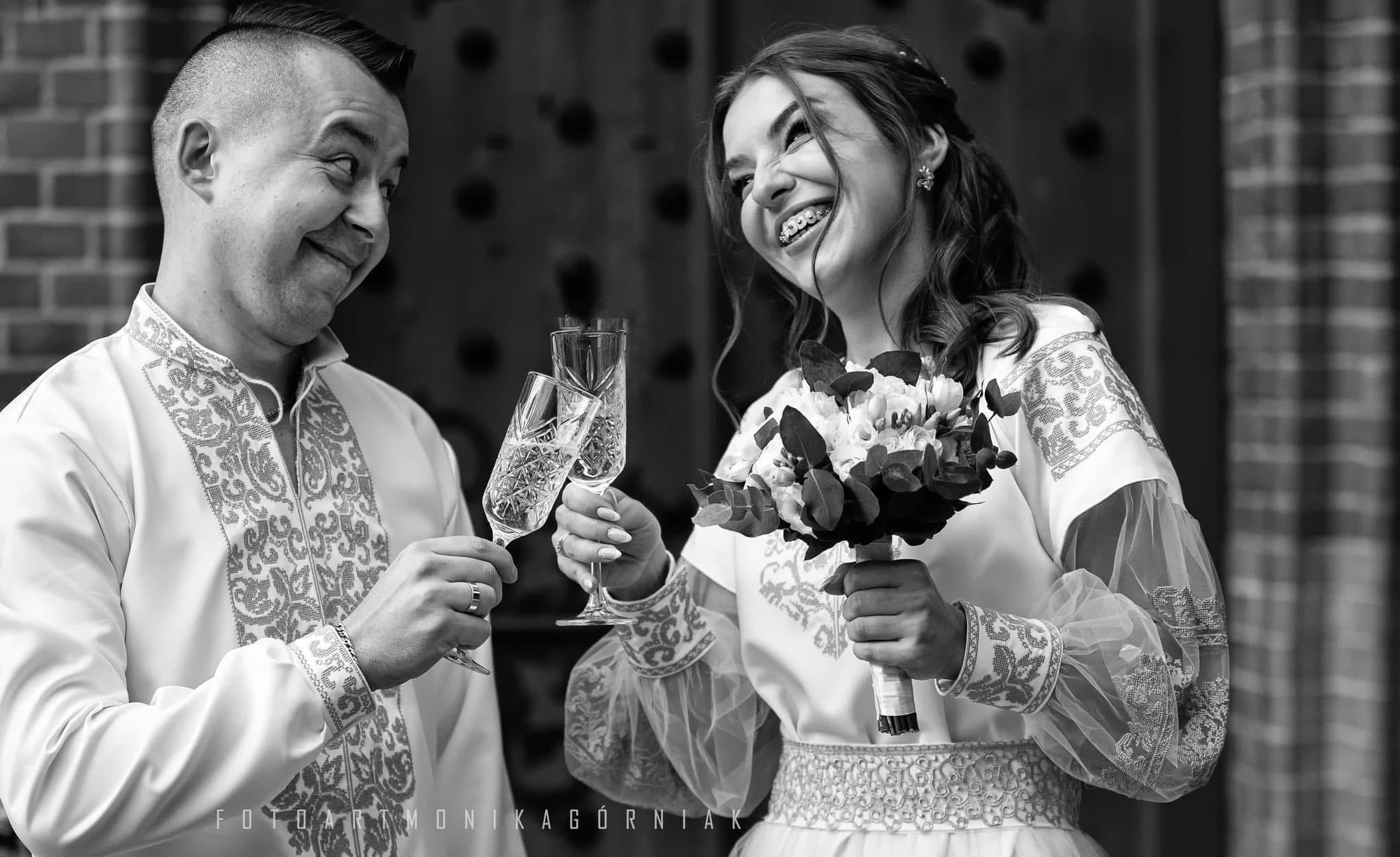 Czarno-białe zdjęcie uśmiechniętej pary młodej w strojach ludowych, wznoszącej toast kryształowymi kieliszkami, panna młoda trzyma bukiet.