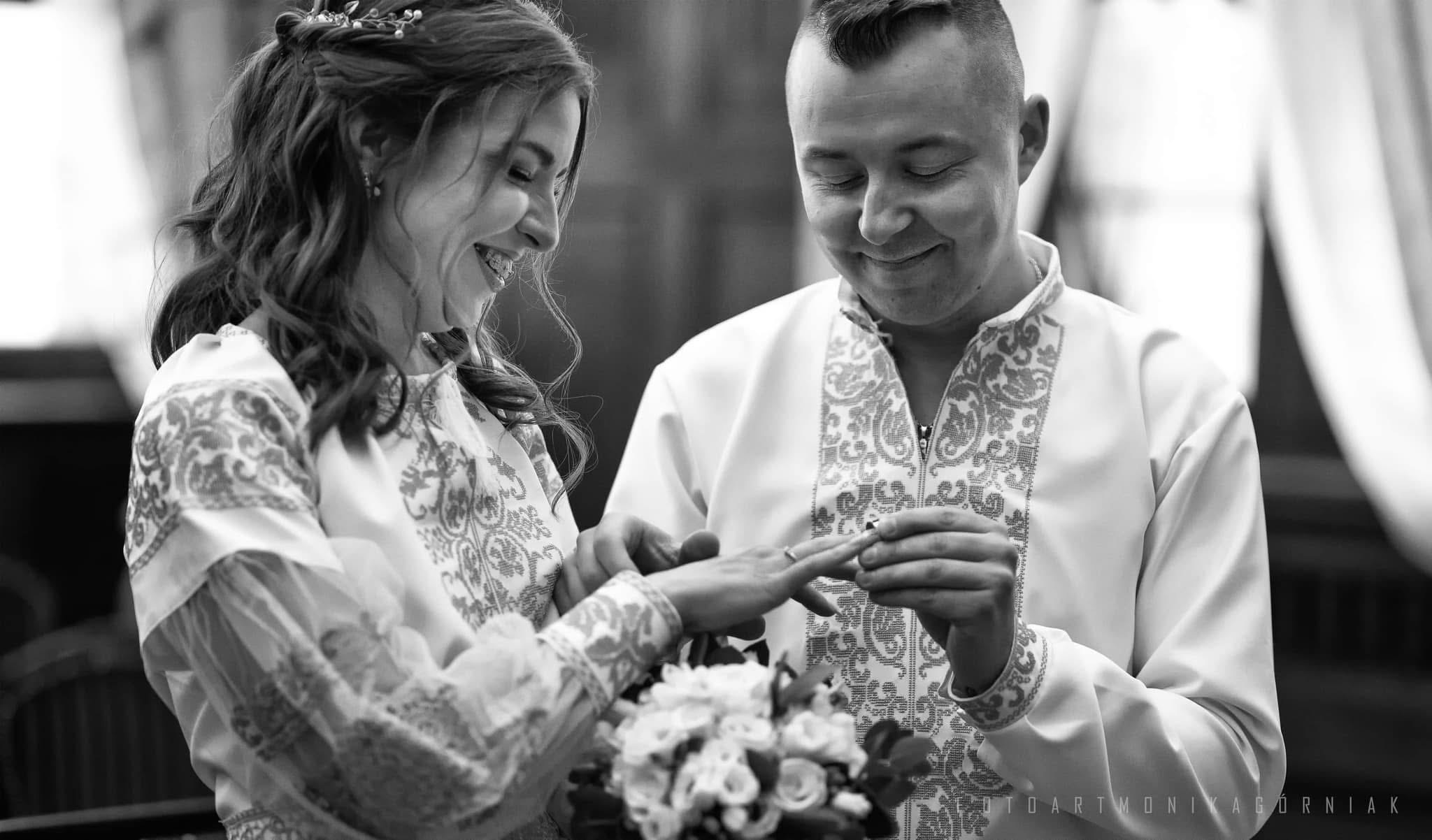 Czarno-białe zdjęcie pary młodej w tradycyjnych strojach podczas ceremonii zakładania obrączki, z bukietem kwiatów. Delikatne oświetlenie podkreśla detale haftu.