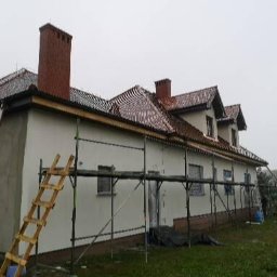 Walldonegk - Dom jednorodzinny w trakcie prac elewacyjnych, widoczne rusztowania, częściowo otynkowana ściana boczna, drewniana drabina oparta o ścianę, dach z czerwonej dachówki, pochmurne niebo.