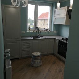 Walldonegk - Jasnoszara kuchnia z drewnianą podłogą, widok na okno z narzędziami, nowoczesne oświetlenie i niebieski taboret malarski.