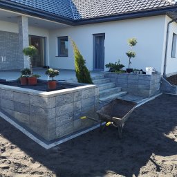 Walldonegk - Nowoczesny dom jednorodzinny z szarą elewacją i kostką brukową, widać elementy architektury krajobrazu: podwyższoną rabatę z roślinami w donicach, schody z kostki i taczkę z ziemią na przygotowanym...