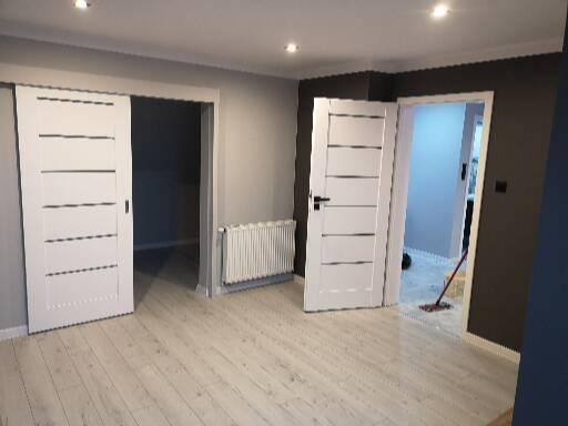 Jasne wnętrze po remoncie z białymi drzwiami z poziomymi panelami, grzejnikiem i podłogą z paneli w odcieniach bieli i szarości, widoczny mop w sąsiednim pokoju.