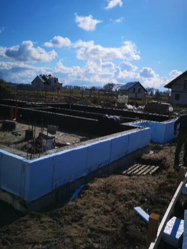 Fundamenty domu w trakcie budowy, ocieplone styropianem, widoczne zbrojenia i szalunki, w tle domy jednorodzinne pod błękitnym niebem z chmurami.