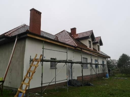 Dom jednorodzinny w trakcie prac elewacyjnych, widoczne rusztowania, częściowo otynkowana ściana boczna, drewniana drabina oparta o ścianę, dach z czerwonej dachówki, pochmurne niebo.