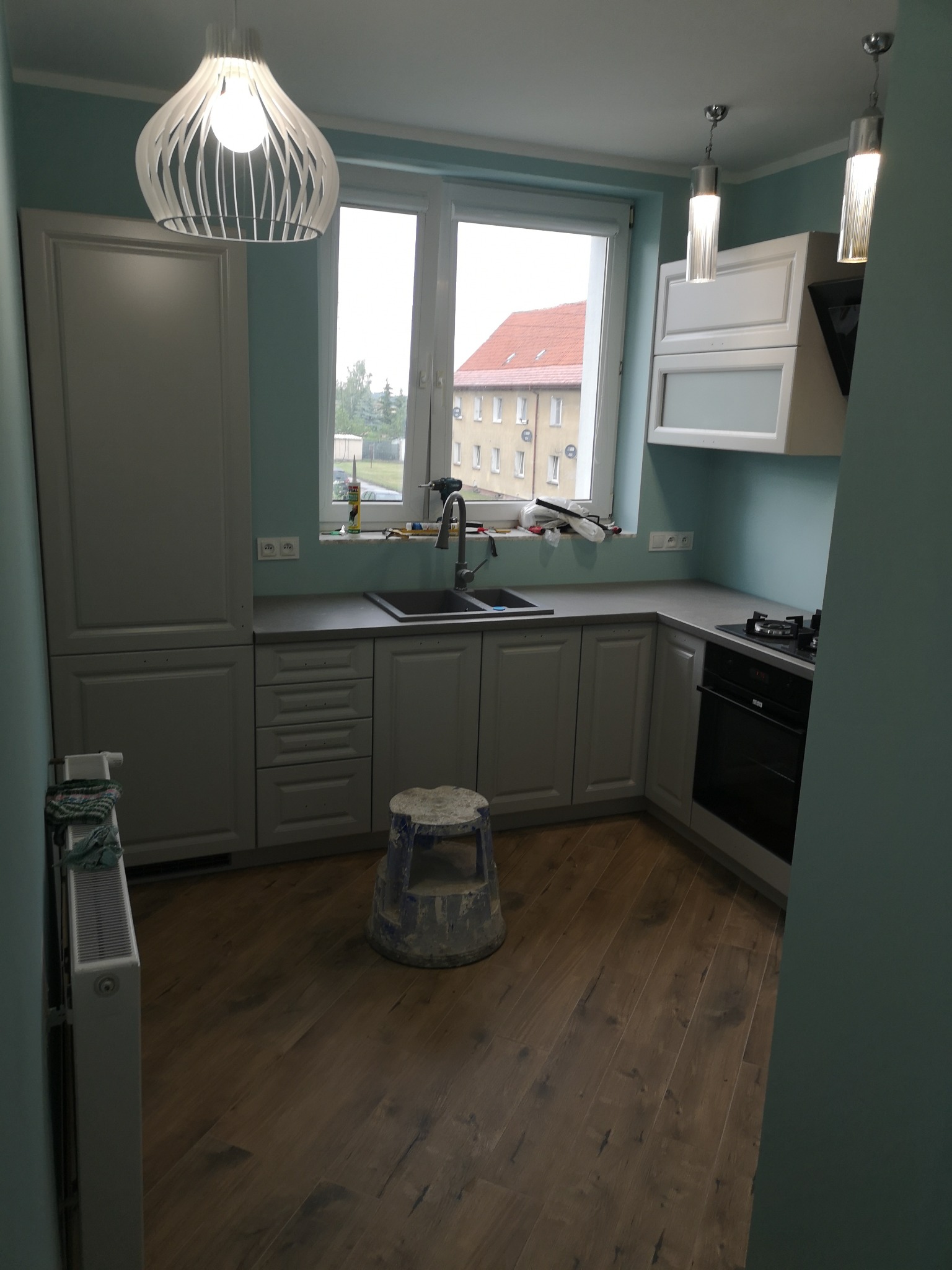 Jasnoszara kuchnia z drewnianą podłogą, widok na okno z narzędziami, nowoczesne oświetlenie i niebieski taboret malarski.