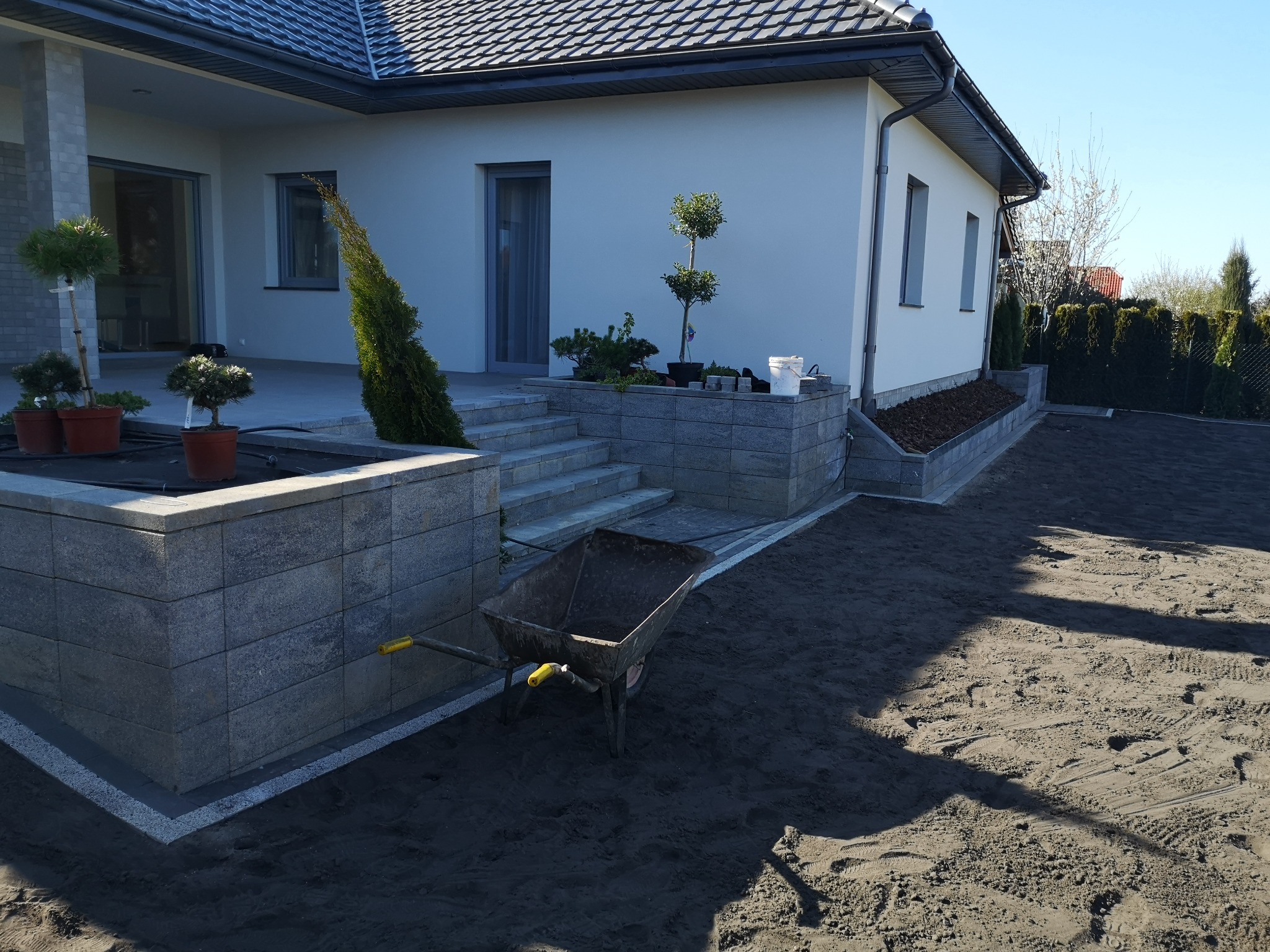 Przygotowanie terenu pod trawnik z użyciem taczki na tle nowoczesnego domu z szarą elewacją i ozdobnymi roślinami.