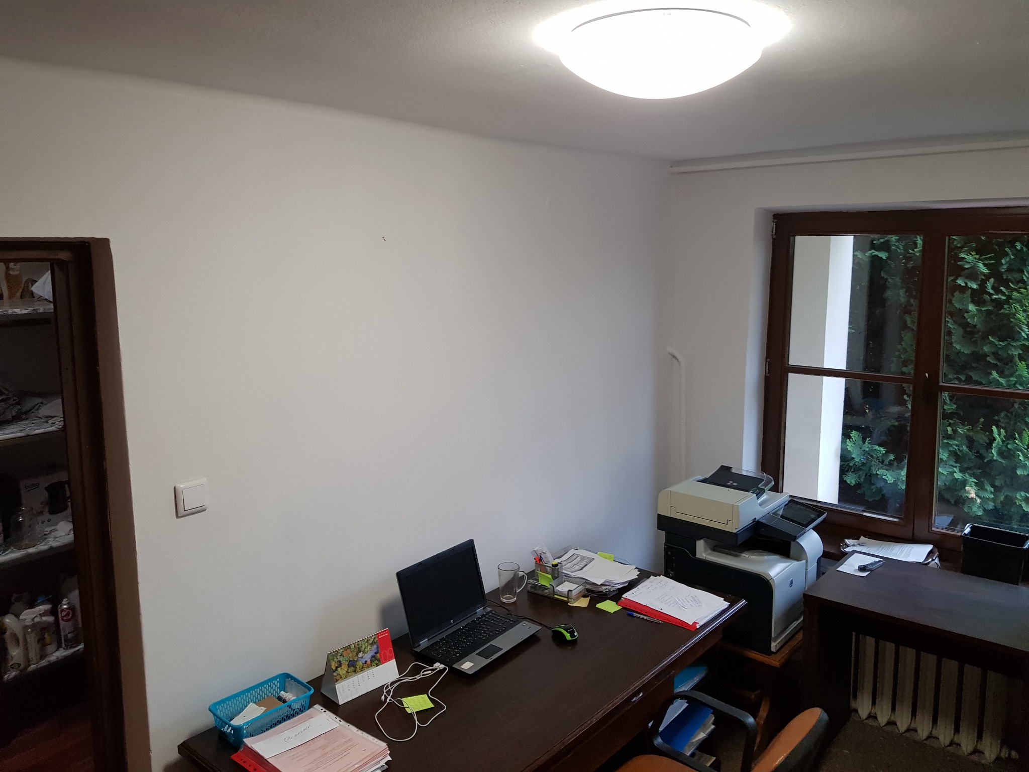 Biała ściana biura przed malowaniem, widoczny włącznik światła, biurko z laptopem, drukarka przy oknie z widokiem na zieleń.