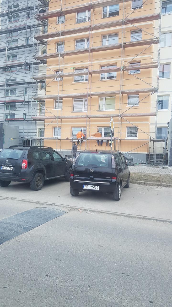 Malowanie elewacji budynku wielorodzinnego w Elblągu, widoczne rusztowania i pracownicy w pomarańczowych kombinezonach, na pierwszym planie zaparkowane samochody.