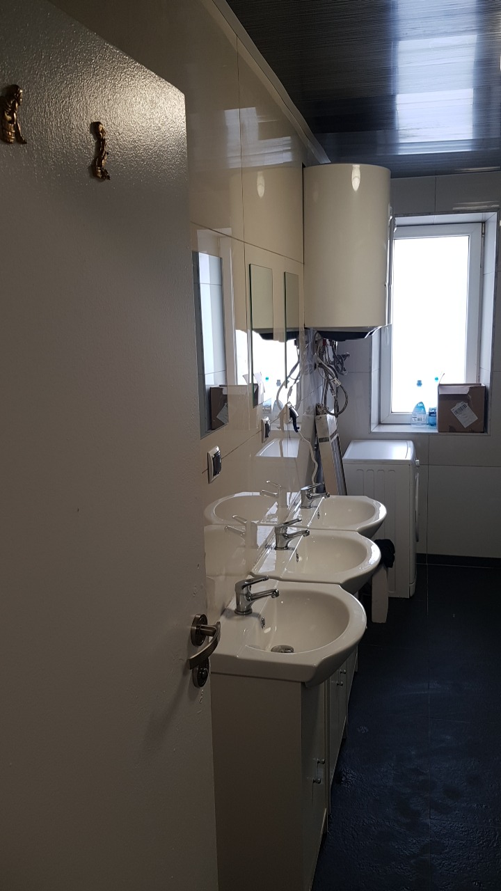 Rząd trzech umywalek z szafkami w białej łazience w Lublinie, z lustrami i bojlerem, widoczna pralka i kartonowe pudło przy oknie.