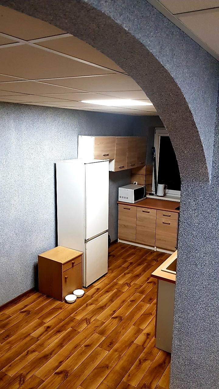 Kuchnia z drewnianą podłogą, białymi sprzętami i szafkami w odcieniach drewna, widok przez szary łuk z fakturą.