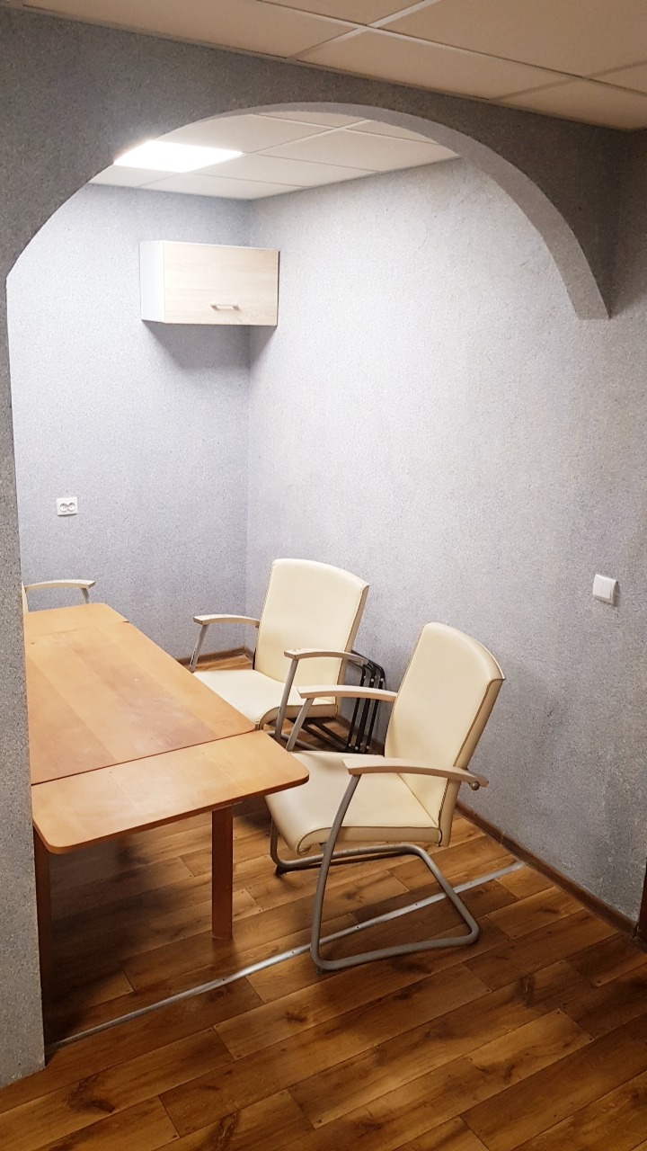 Biuro z drewnianym stołem i krzesłami z kremową tapicerką, wykończone ścianami o szarej, ziarnistej fakturze i łukowatym przejściem.