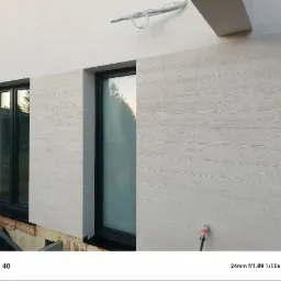 Elewacja budynku z oknami, wykończona panelami imitującymi drewno w jasnym odcieniu. Widoczne detale wykończenia i instalacja na ścianie.