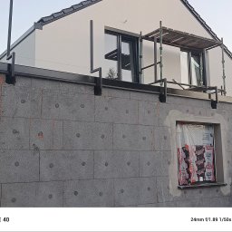 Firma ogólno-budowlana PawBud - Szara elewacja w trakcie ocieplania, widoczne płyty izolacyjne z charakterystycznymi wgłębieniami, okno zabezpieczone folią, fragment rusztowania i konstrukcji dachu.